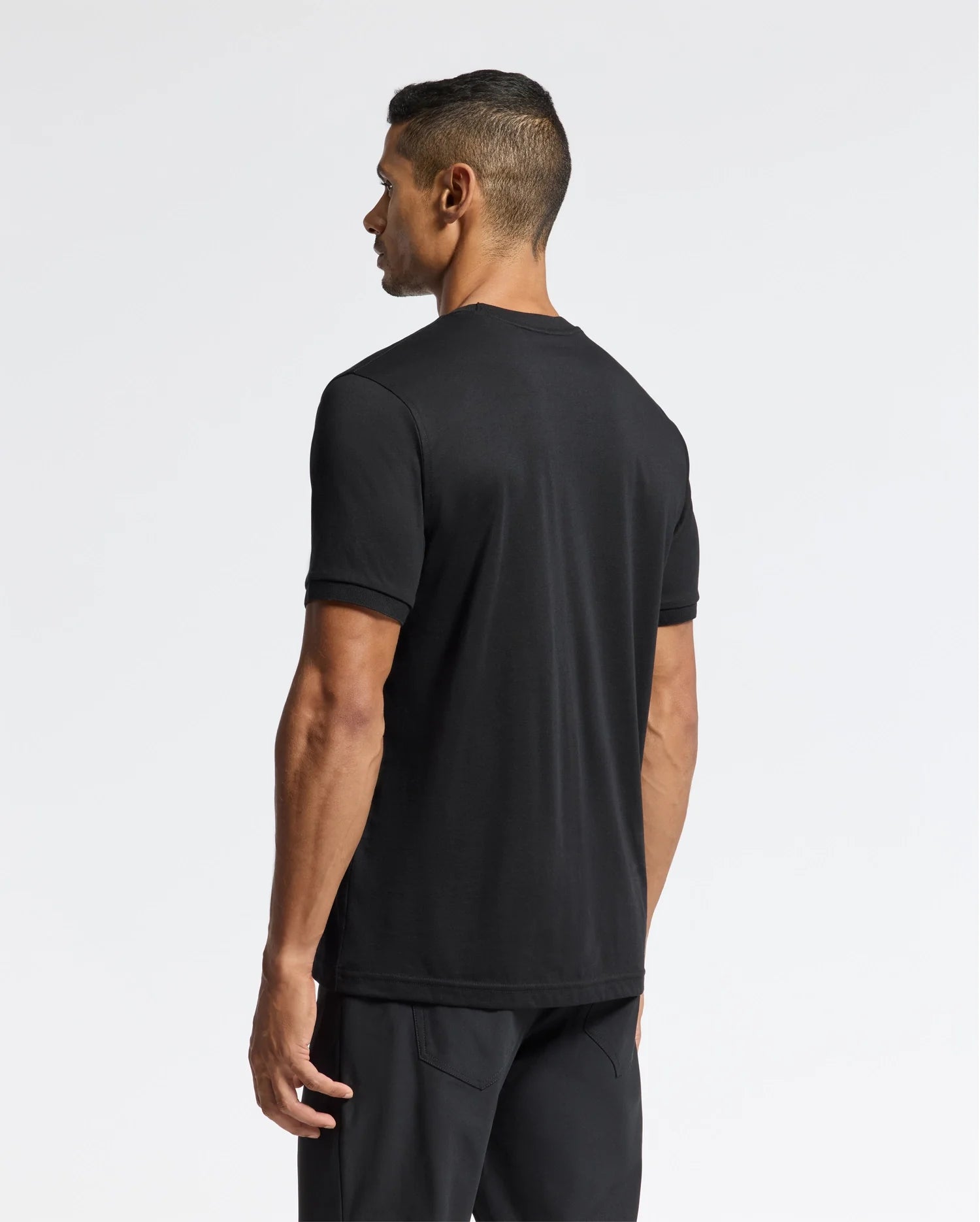 A man with short black hair stands facing away from the camera, dressed in the premium quality Psycho Bunny MENS BRANSON FASHION TEE - B6U137E200 and black pants against a plain white background.