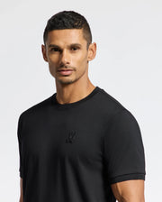 A man dressed in a Psycho Bunny MENS BRANSON FASHION TEE - B6U137E200, featuring a premium embroidered logo on black Pima cotton, stands against a light gray backdrop. This lightweight fashion piece highlights modern men's style. He sports short dark hair and a light beard while gazing directly at the camera.