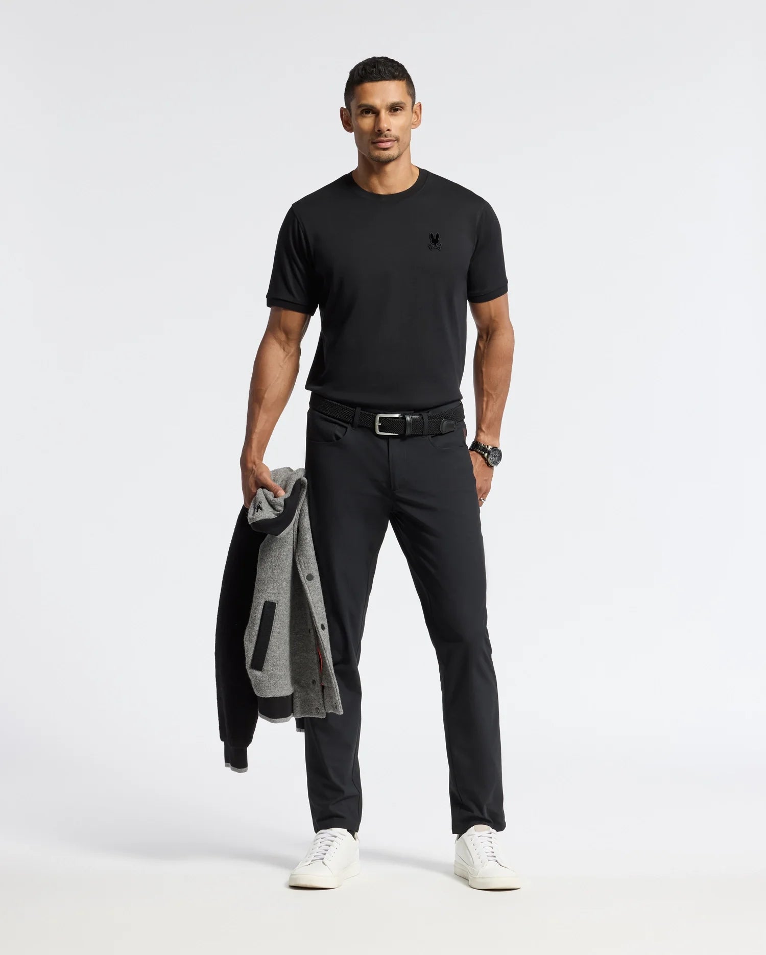 A man, dressed in Psycho Bunny's black MENS BRANSON FASHION TEE - B6U137E200 and pants, holds a gray jacket. Against a plain white background, he's accessorized with a black belt and watch, perfectly complemented by white sneakers.
