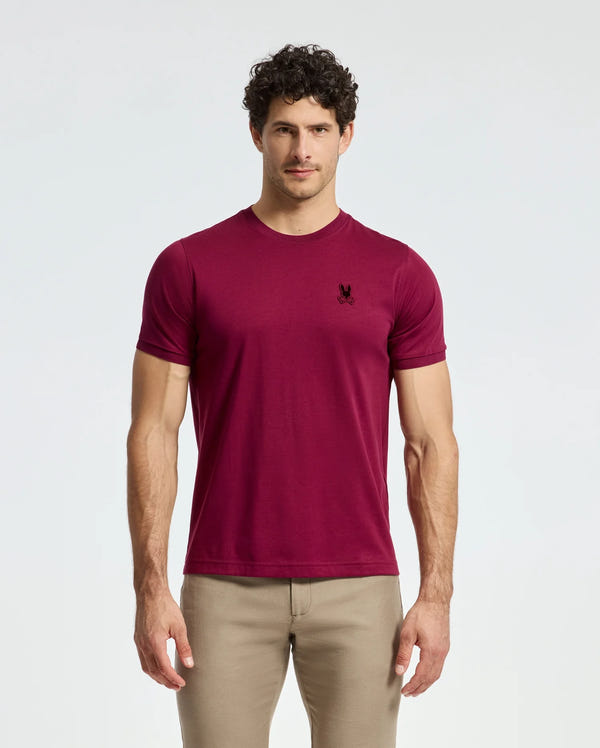 A man with curly hair stands against a plain background, wearing a fashion-forward Psycho Bunny MENS BRANSON FASHION TEE - B6U137E200 in burgundy Pima cotton with an embroidered emblem on the chest and beige pants.