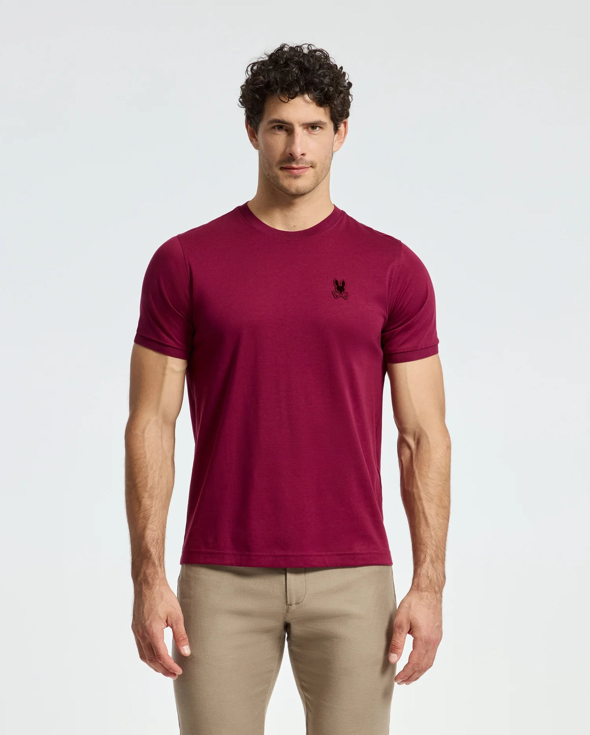A man with curly hair stands against a plain background, wearing a fashion-forward Psycho Bunny MENS BRANSON FASHION TEE - B6U137E200 in burgundy Pima cotton with an embroidered emblem on the chest and beige pants.