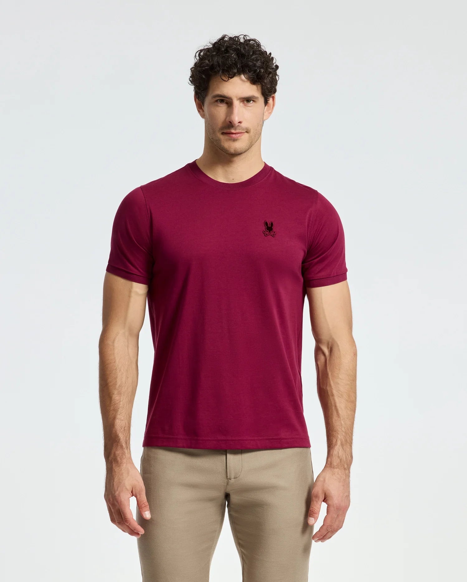 A man with curly hair stands against a plain background, wearing a fashion-forward Psycho Bunny MENS BRANSON FASHION TEE - B6U137E200 in burgundy Pima cotton with an embroidered emblem on the chest and beige pants.