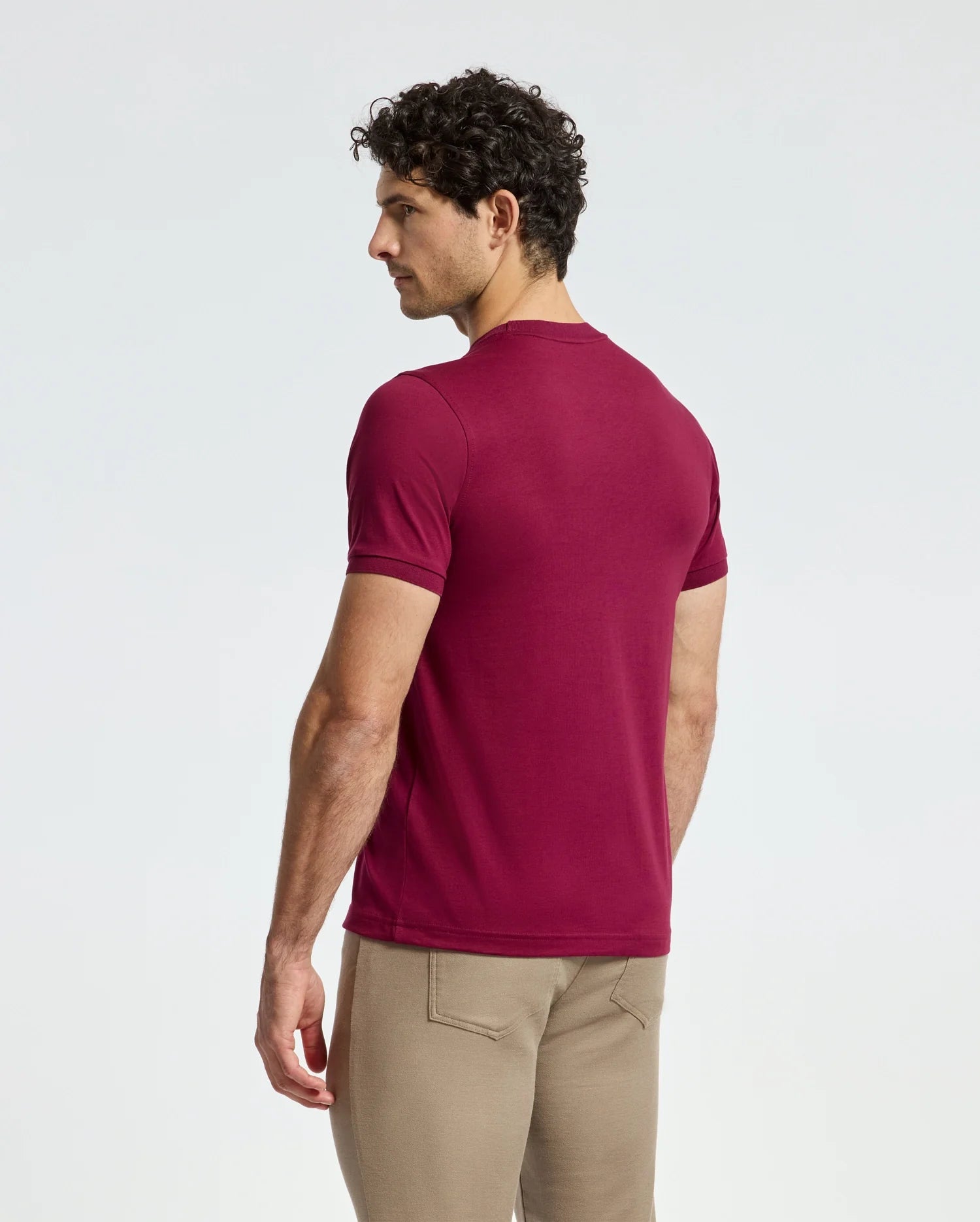 A man with curly hair, facing away, is dressed in a fashion-forward Psycho Bunny MENS BRANSON FASHION TEE - B6U137E200 and beige pants. The plain white background accentuates the tee's sleek, lightweight design.