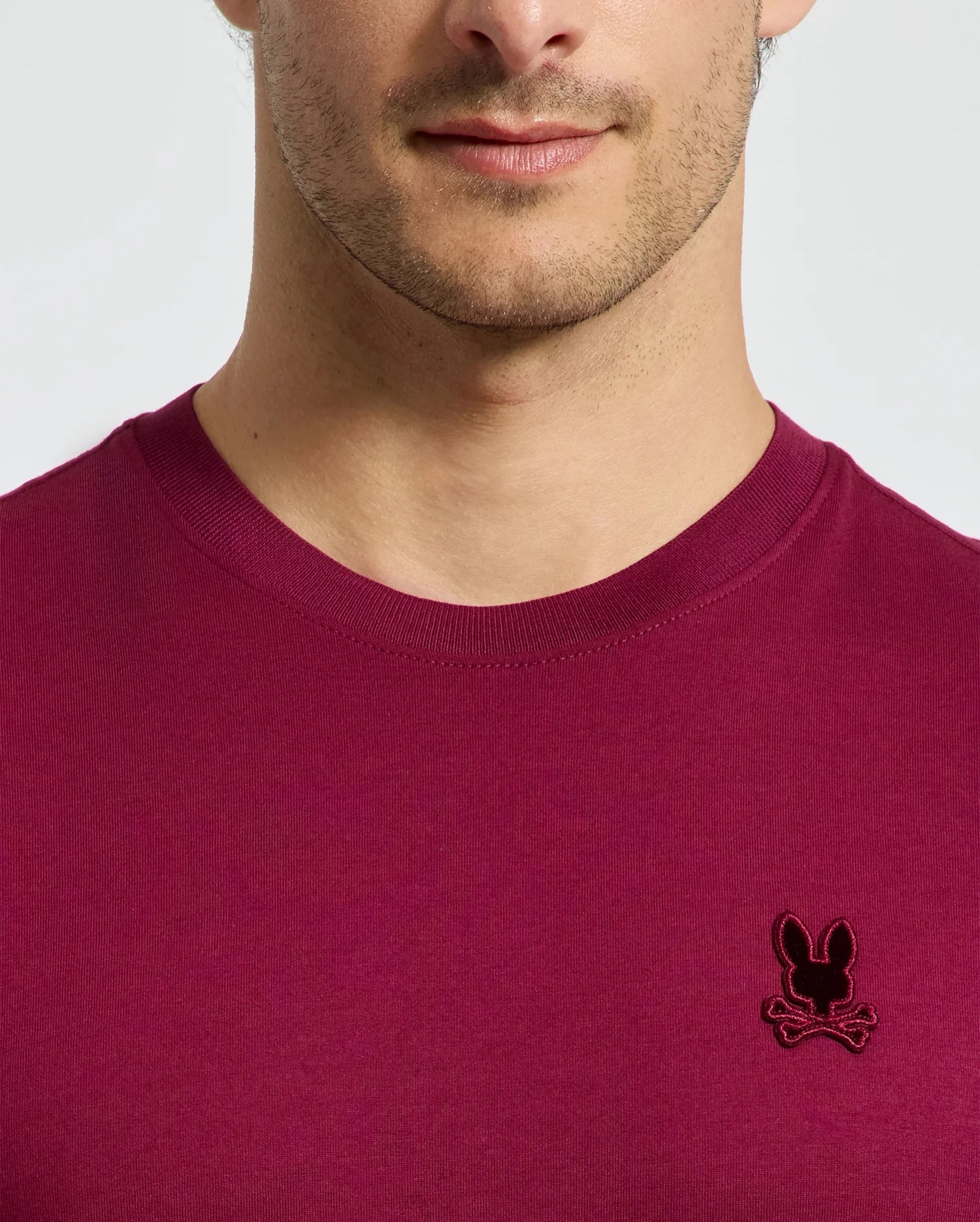 A person is seen in a stylish Psycho Bunny MENS BRANSON FASHION TEE - B6U137E200, crafted from lightweight cotton in a vibrant magenta color. The tee features a distinctive black embroidered bunny logo on the chest. The lower half of the face and upper torso are visible against a plain background, highlighting the tee's chic and effortless style.