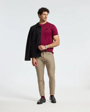 A man stands confidently wearing a fashion-forward burgundy "MENS BRANSON FASHION TEE - B6U137E200" by Psycho Bunny, paired with beige pants and black loafers. He holds a black jacket over his shoulder while gazing slightly to the left, with his other hand in his pocket. The background is plain white.