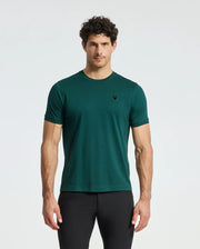 A man with curly hair stands against a plain background, wearing a dark green Psycho Bunny Men's Branson Fashion Tee - B6U137E200, featuring a small black logo on the left chest and paired with black pants.