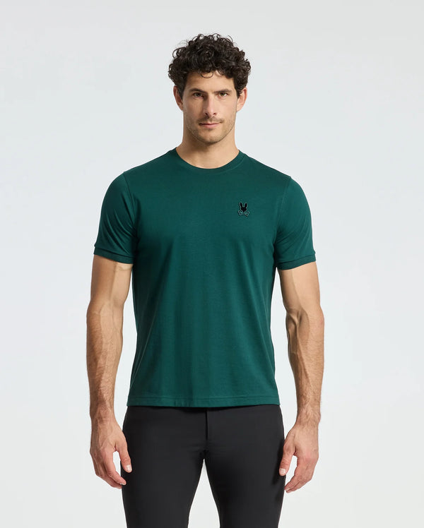 A man with curly hair stands against a plain background, wearing a dark green Psycho Bunny Men's Branson Fashion Tee - B6U137E200, featuring a small black logo on the left chest and paired with black pants.