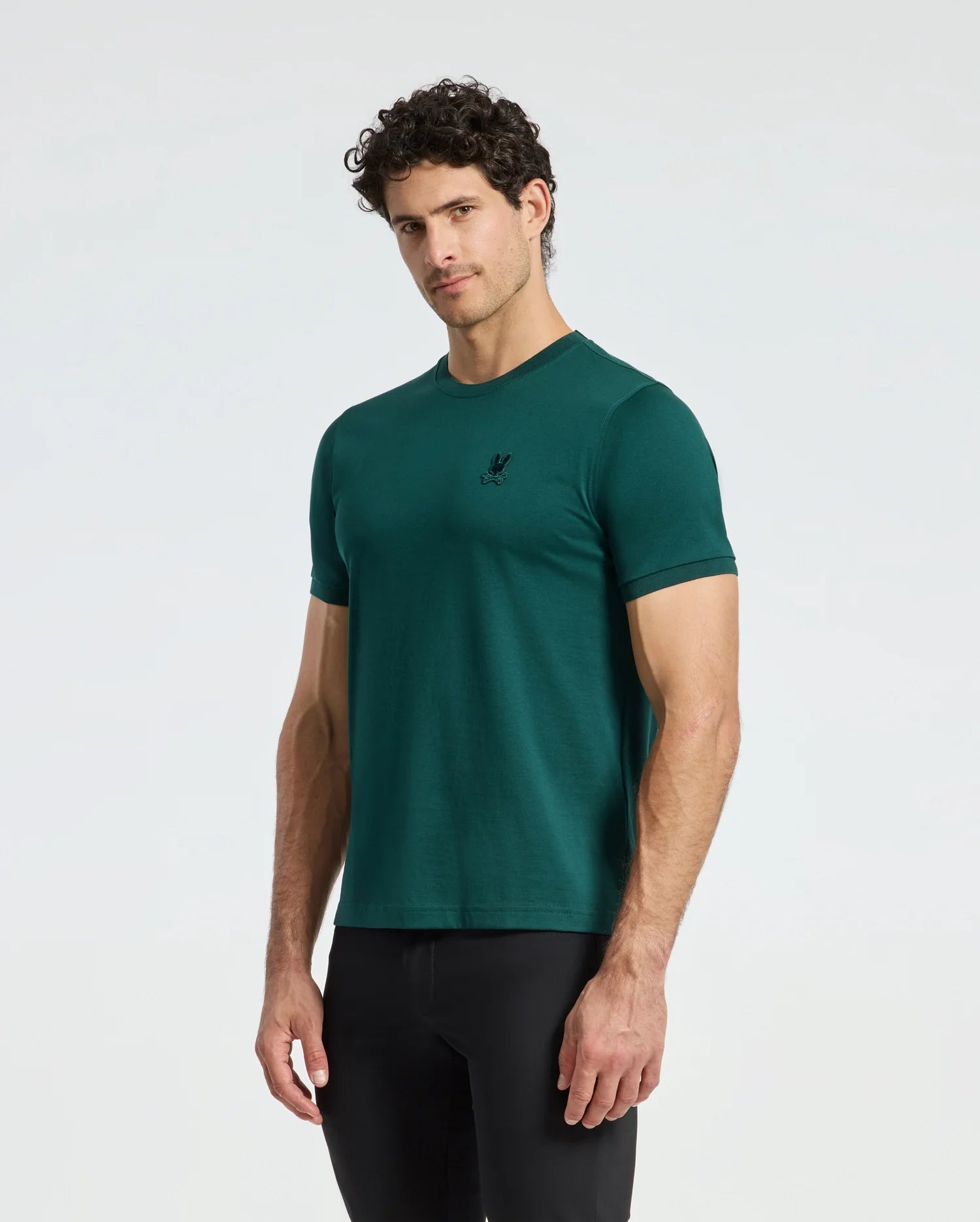 A man with curly hair is wearing a green Psycho Bunny MENS BRANSON FASHION TEE - B6U137E200 and black pants, standing against a plain white background. The tee, renowned for its lightweight comfort, features a small logo on the chest.