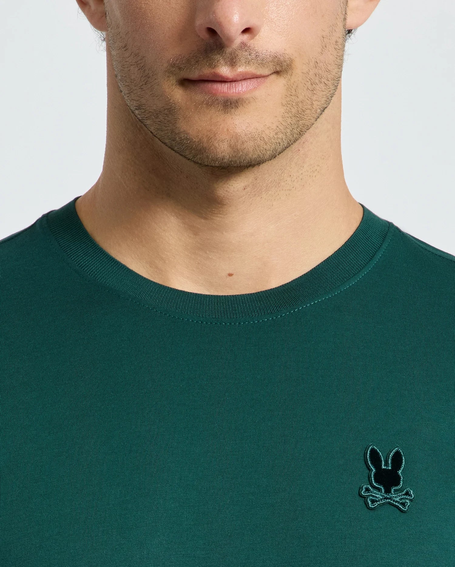 A person wearing a dark green Psycho Bunny MENS BRANSON FASHION TEE - B6U137E200, featuring a small embroidered emblem of a bunny and crossbones on the chest. The focus is on the shirt's lightweight fabric and design, rather than the person's face.