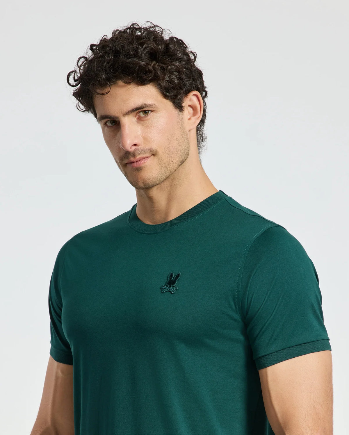 A man with short curly hair wears a dark green Psycho Bunny MENS BRANSON FASHION TEE (B6U137E200) that features a small embroidered logo on the chest. He stands against a plain white background, looking slightly to the side.