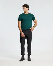 A man wearing a Men's Branson Fashion Tee from Psycho Bunny, paired with black pants and black boots, stands against a plain white background. He is looking to the side with a casual posture and is accessorized with a gold necklace and bracelet.