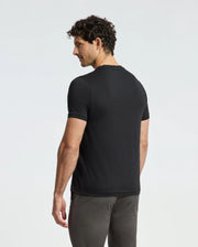 A man with curly hair stands facing away, effortlessly showcasing the premium quality of the MENS ALEXANDER FASHION TEE - B6U139E200 by Psycho Bunny. Dressed in a stylish black tee and gray pants, he looks over his shoulder against a plain white background.