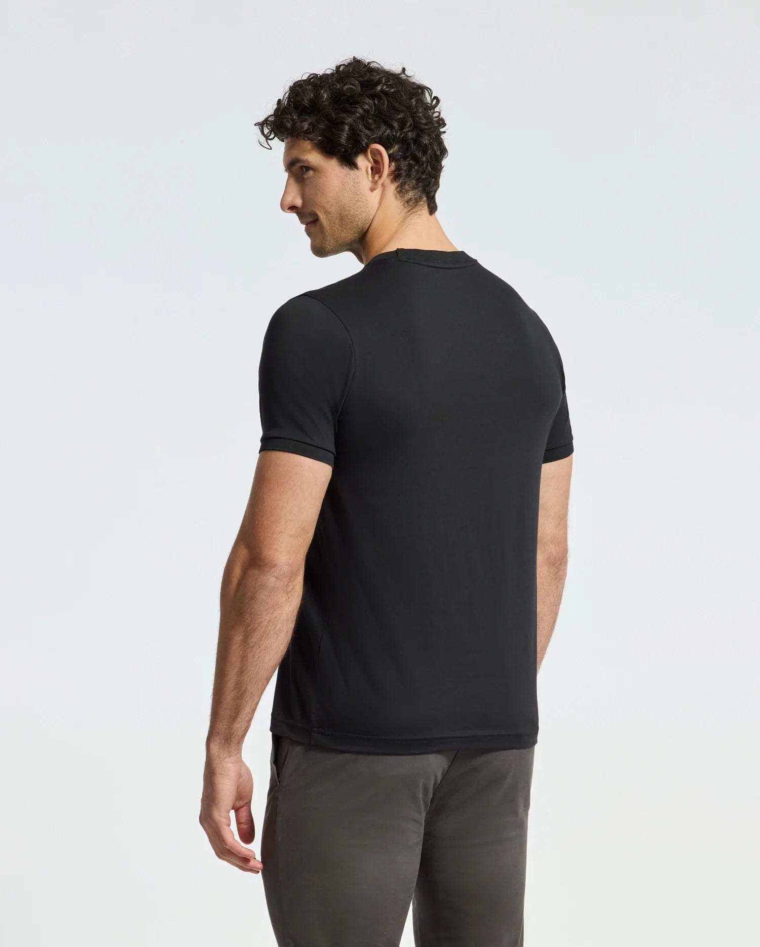 A man with curly hair stands facing away, effortlessly showcasing the premium quality of the MENS ALEXANDER FASHION TEE - B6U139E200 by Psycho Bunny. Dressed in a stylish black tee and gray pants, he looks over his shoulder against a plain white background.