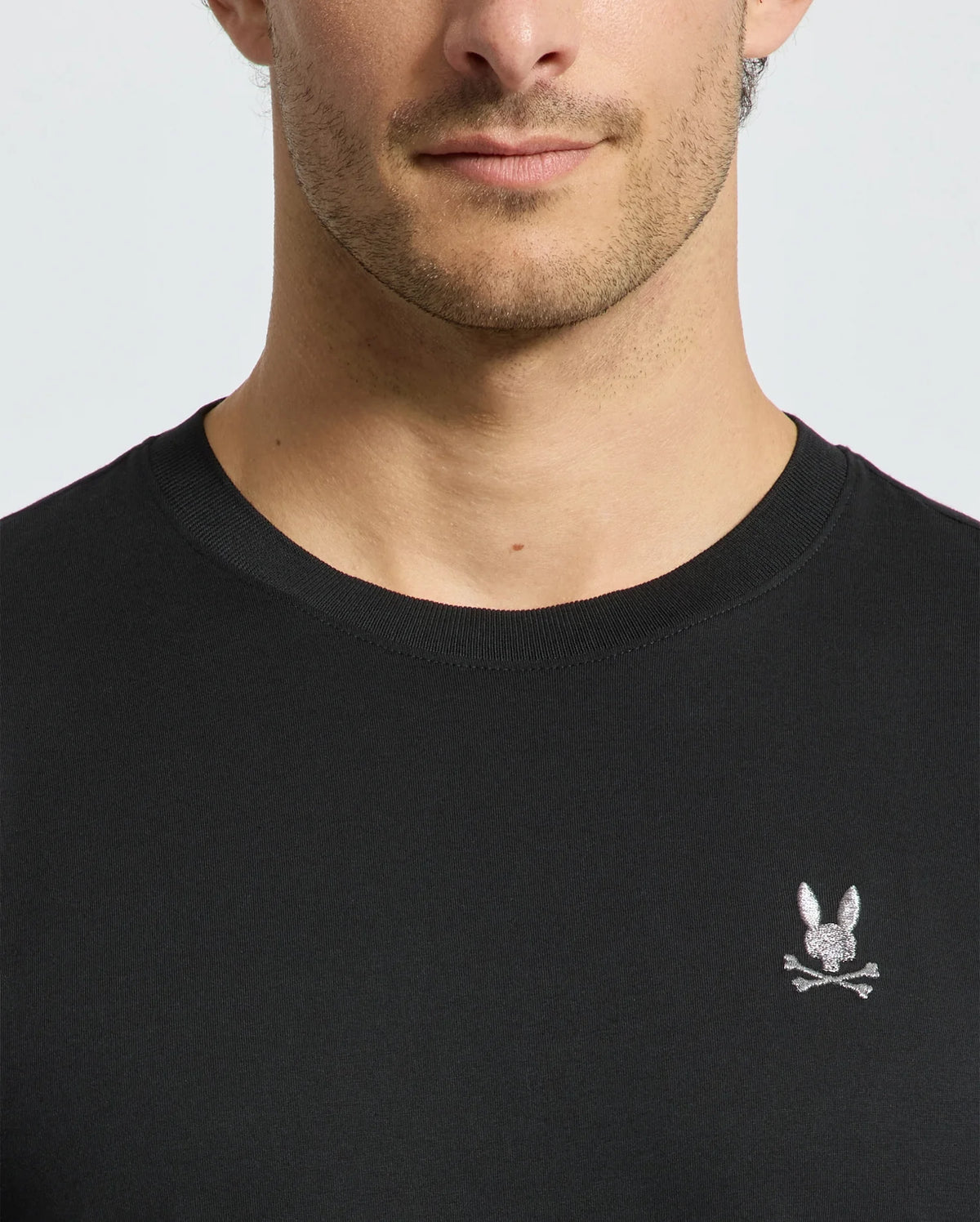 A close-up image captures a man wearing the luxurious Psycho Bunny MENS ALEXANDER FASHION TEE - B6U139E200 in black, which is adorned with a subtle embroidered rabbit and crossbones design on the chest. His face is partially visible from the nose down, highlighting the premium quality of this Pima cotton T-shirt.