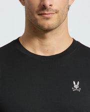 A close-up image captures a man wearing the luxurious Psycho Bunny MENS ALEXANDER FASHION TEE - B6U139E200 in black, which is adorned with a subtle embroidered rabbit and crossbones design on the chest. His face is partially visible from the nose down, highlighting the premium quality of this Pima cotton T-shirt.