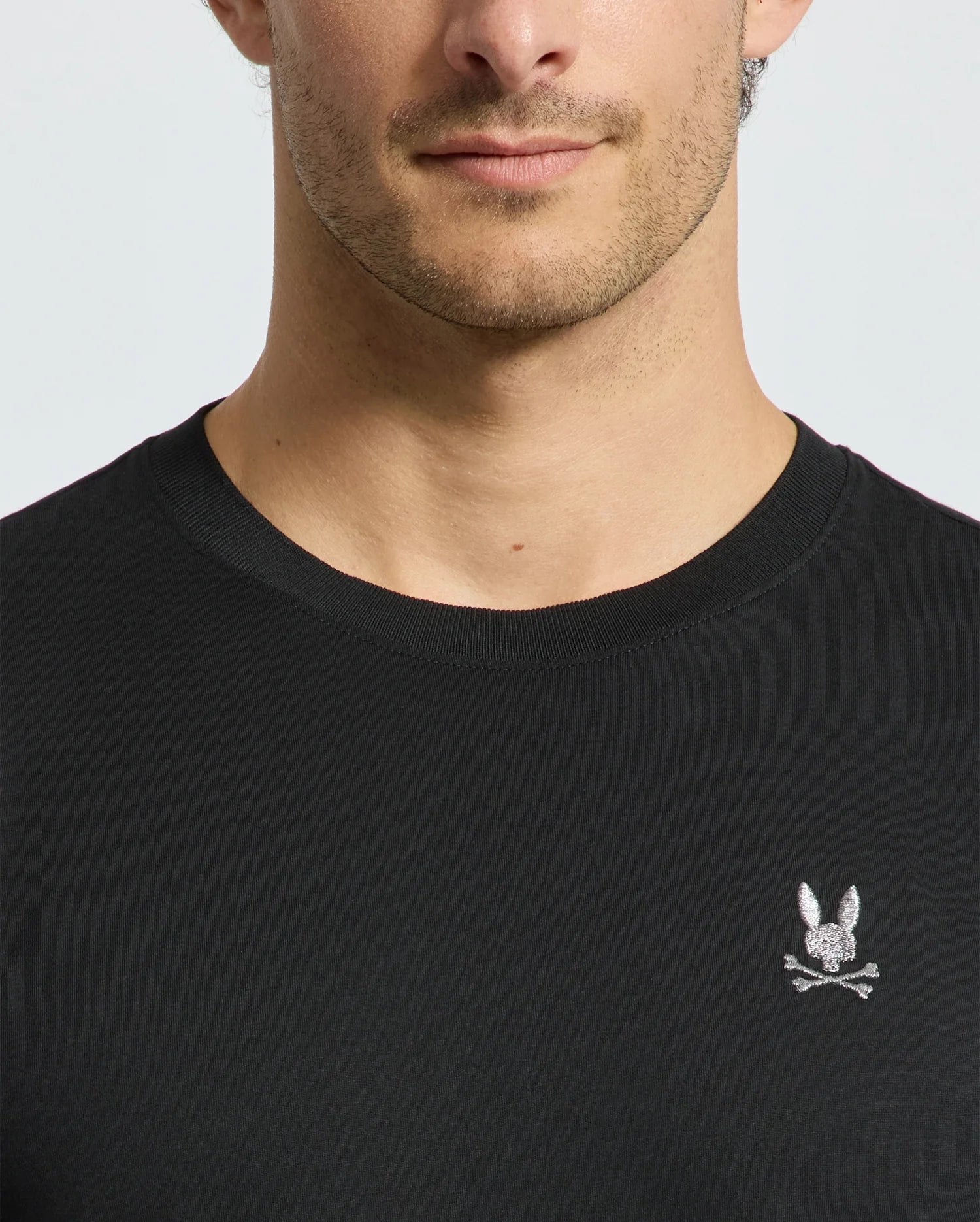 A close-up image captures a man wearing the luxurious Psycho Bunny MENS ALEXANDER FASHION TEE - B6U139E200 in black, which is adorned with a subtle embroidered rabbit and crossbones design on the chest. His face is partially visible from the nose down, highlighting the premium quality of this Pima cotton T-shirt.