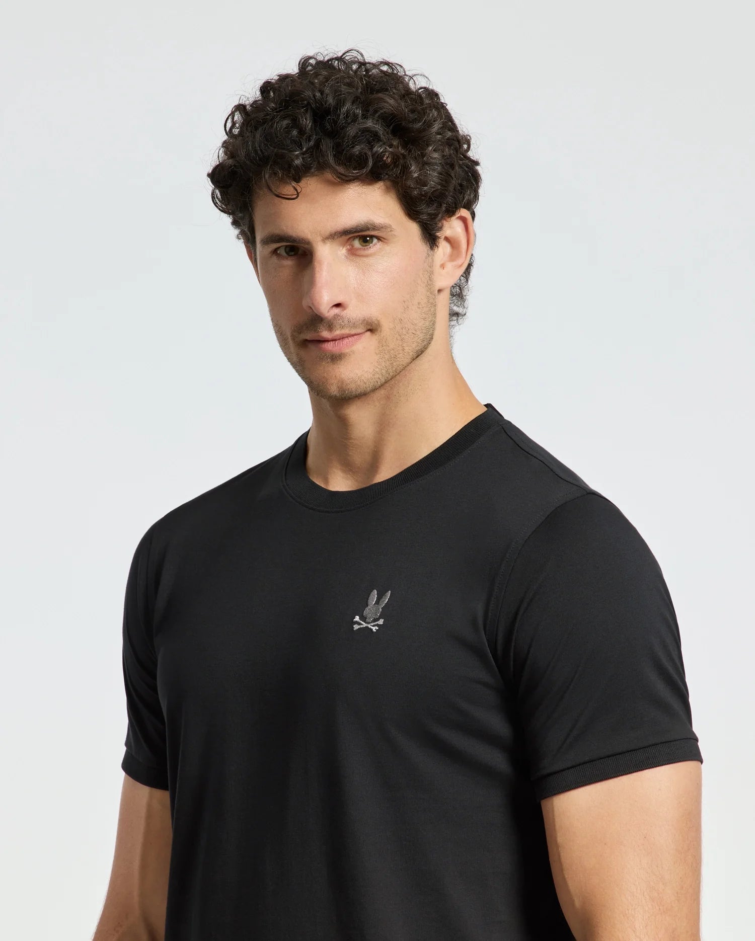 A man with curly hair is wearing the Psycho Bunny MENS ALEXANDER FASHION TEE - B6U139E200, showcasing a small embroidered eagle logo on the chest. He stands against a plain white background, looking slightly towards the camera.
