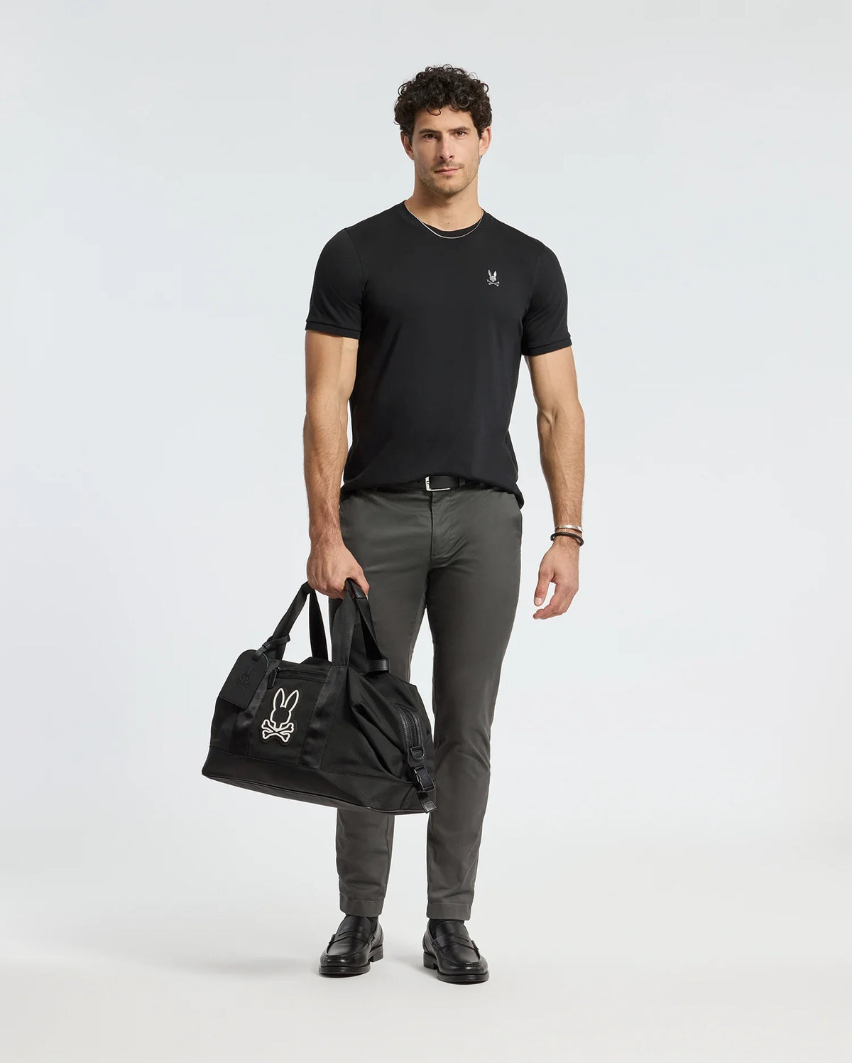 A man in a MENS ALEXANDER FASHION TEE - B6U139E200 by Psycho Bunny and gray pants stands holding a large black bag adorned with a white logo. He is wearing black shoes and a bracelet. The plain white background complements his premium quality style.