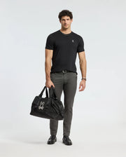A man in a MENS ALEXANDER FASHION TEE - B6U139E200 by Psycho Bunny and gray pants stands holding a large black bag adorned with a white logo. He is wearing black shoes and a bracelet. The plain white background complements his premium quality style.