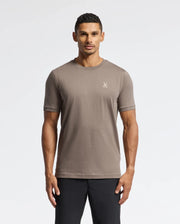 A man with short dark hair is wearing the luxurious Psycho Bunny MENS ALEXANDER FASHION TEE - B6U139E200 in taupe, which features a small chest logo. He pairs it with black pants and stands against a plain white background, facing forward with his arms relaxed at his sides.