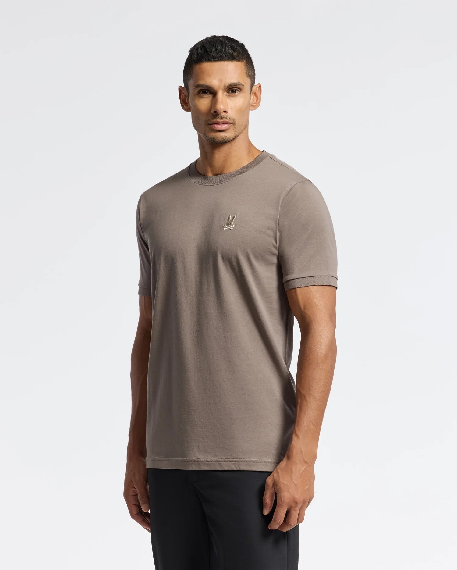 A man wearing a luxurious Psycho Bunny Men's Alexander Fashion Tee (B6U139E200), featuring a small embroidered logo on the chest, and black pants stands in a well-lit studio setting. He has short dark hair and looks directly at the camera. The plain white background highlights the premium quality of this men's fashion T-shirt.