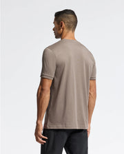 A man stands with his back to the camera, dressed in a taupe-colored Psycho Bunny MENS ALEXANDER FASHION TEE - B6U139E200 and black pants. The white background highlights the clean, minimalist style.