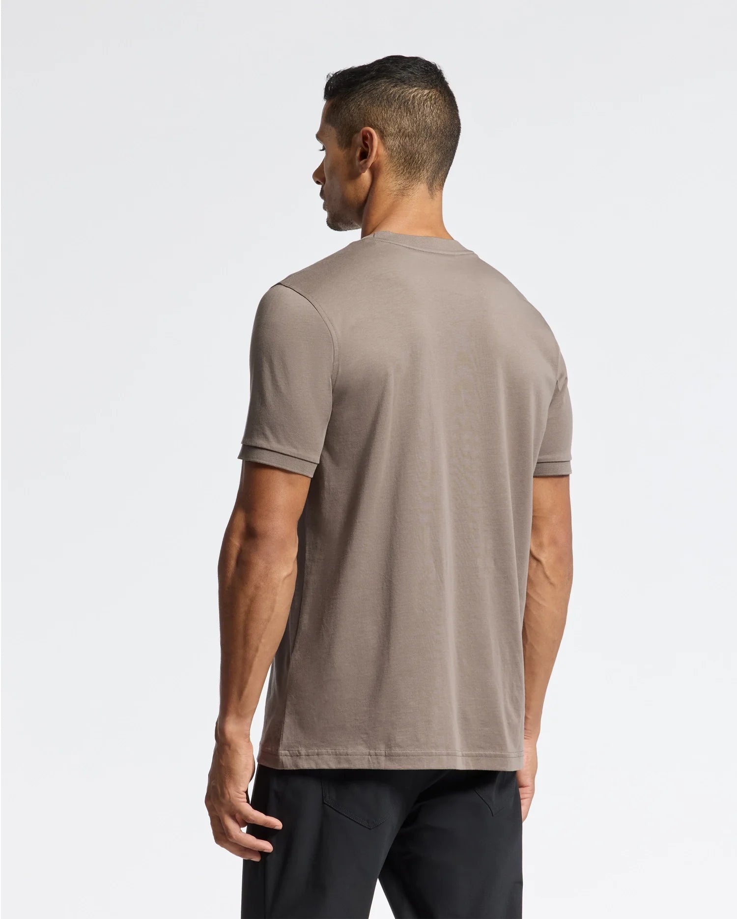 A man stands with his back to the camera, dressed in a taupe-colored Psycho Bunny MENS ALEXANDER FASHION TEE - B6U139E200 and black pants. The white background highlights the clean, minimalist style.
