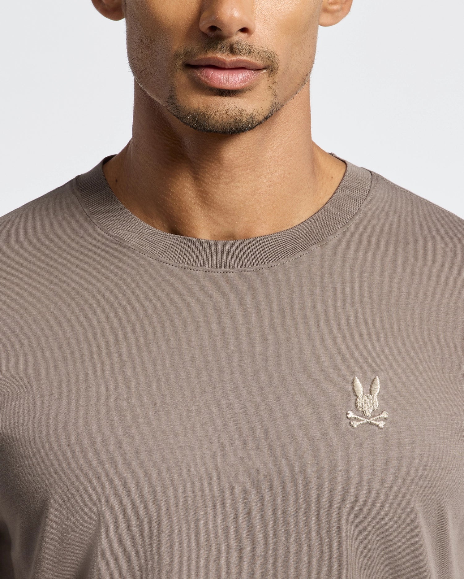A person is wearing the taupe-colored MENS ALEXANDER FASHION TEE from Psycho Bunny. The tee features an embroidered emblem of a rabbit skull and crossbones. The image captures the upper torso and shoulders, emphasizing the premium round neckline of this fashionable piece.