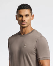 A person dressed in the luxurious MENS ALEXANDER FASHION TEE by Psycho Bunny, featuring an embroidered logo, stands against a plain white background, gazing toward the camera.