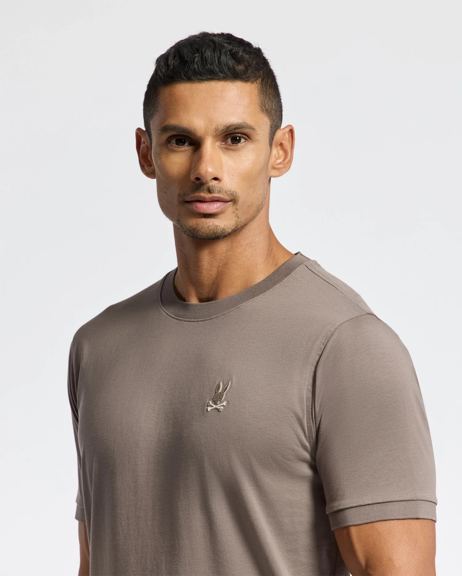 A person dressed in the luxurious MENS ALEXANDER FASHION TEE by Psycho Bunny, featuring an embroidered logo, stands against a plain white background, gazing toward the camera.