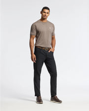 A man stands confidently wearing a luxurious Psycho Bunny MENS ALEXANDER FASHION TEE - B6U139E200 in taupe, paired with black pants and brown shoes. His left hand rests casually in his pocket against a plain white background.