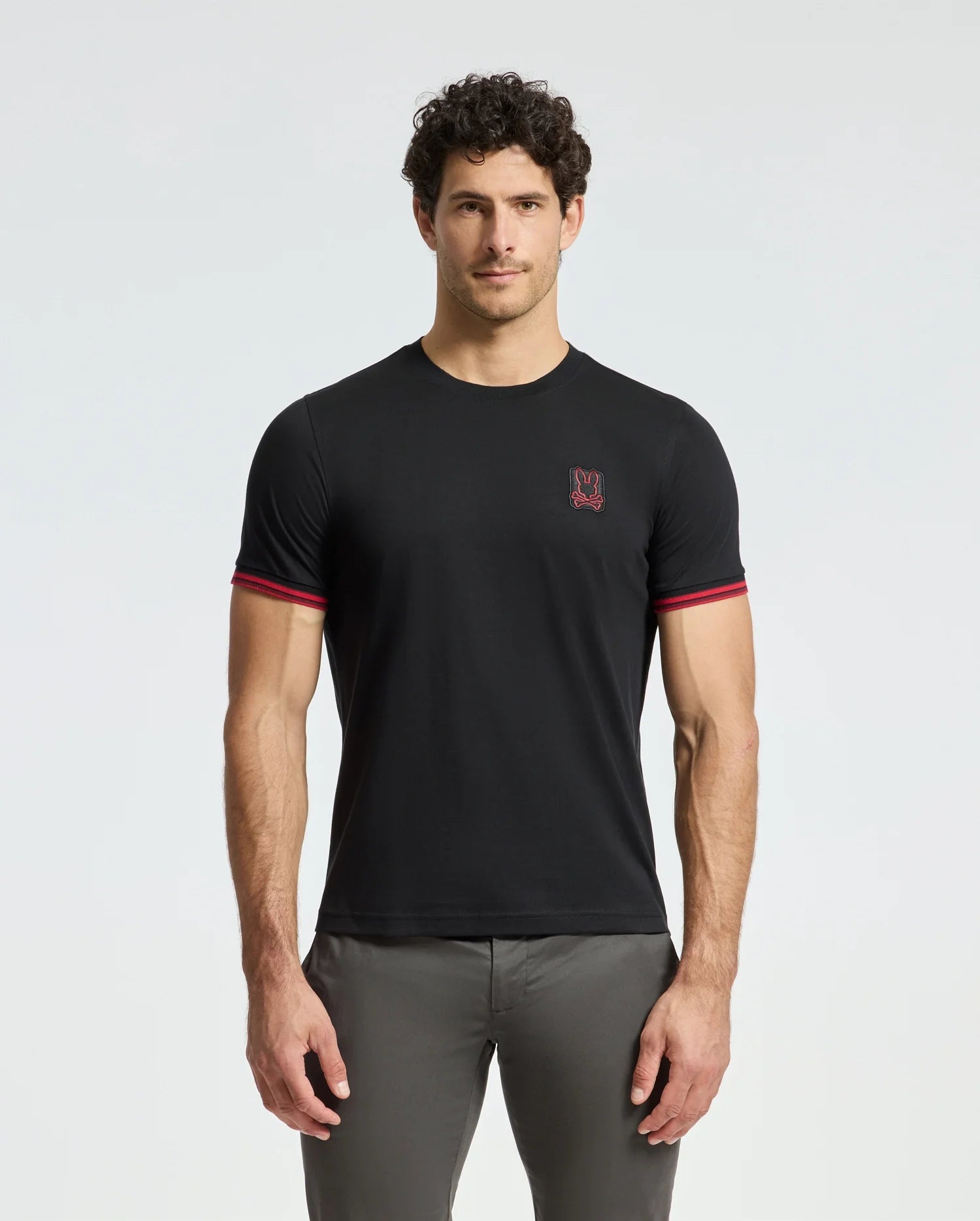 A man with curly hair is wearing the Psycho Bunny MENS PIERCE FASHION TEE (B6U140E200), a black Pima cotton tee featuring red trim on the sleeves and an embroidered Bunny patch on the chest. He stands facing forward with his hands at his sides against a plain light background.