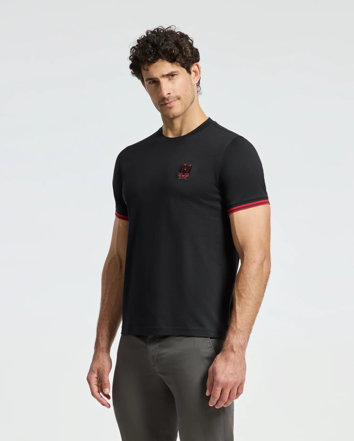 A man with curly hair is wearing the Psycho Bunny MENS PIERCE FASHION TEE in black, featuring red trim on the sleeves and an embroidered Bunny patch on the chest, complemented by gray pants. The luxurious feel of the Pima cotton fabric stands out against the plain white background.
