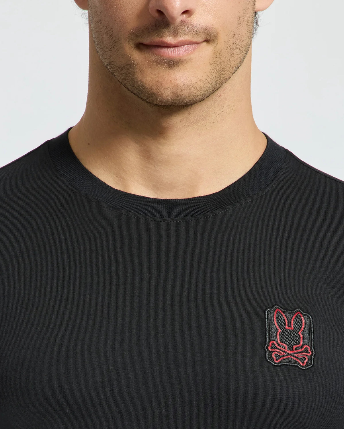 A model wears the Psycho Bunny Men's Pierce Fashion Tee (B6U140E200), made from soft Pima cotton and highlighting an embroidered Bunny patch with crossed bones on the chest. This black T-shirt features a design that is both subtle and stylish, displaying only the lower half of the face and upper torso.