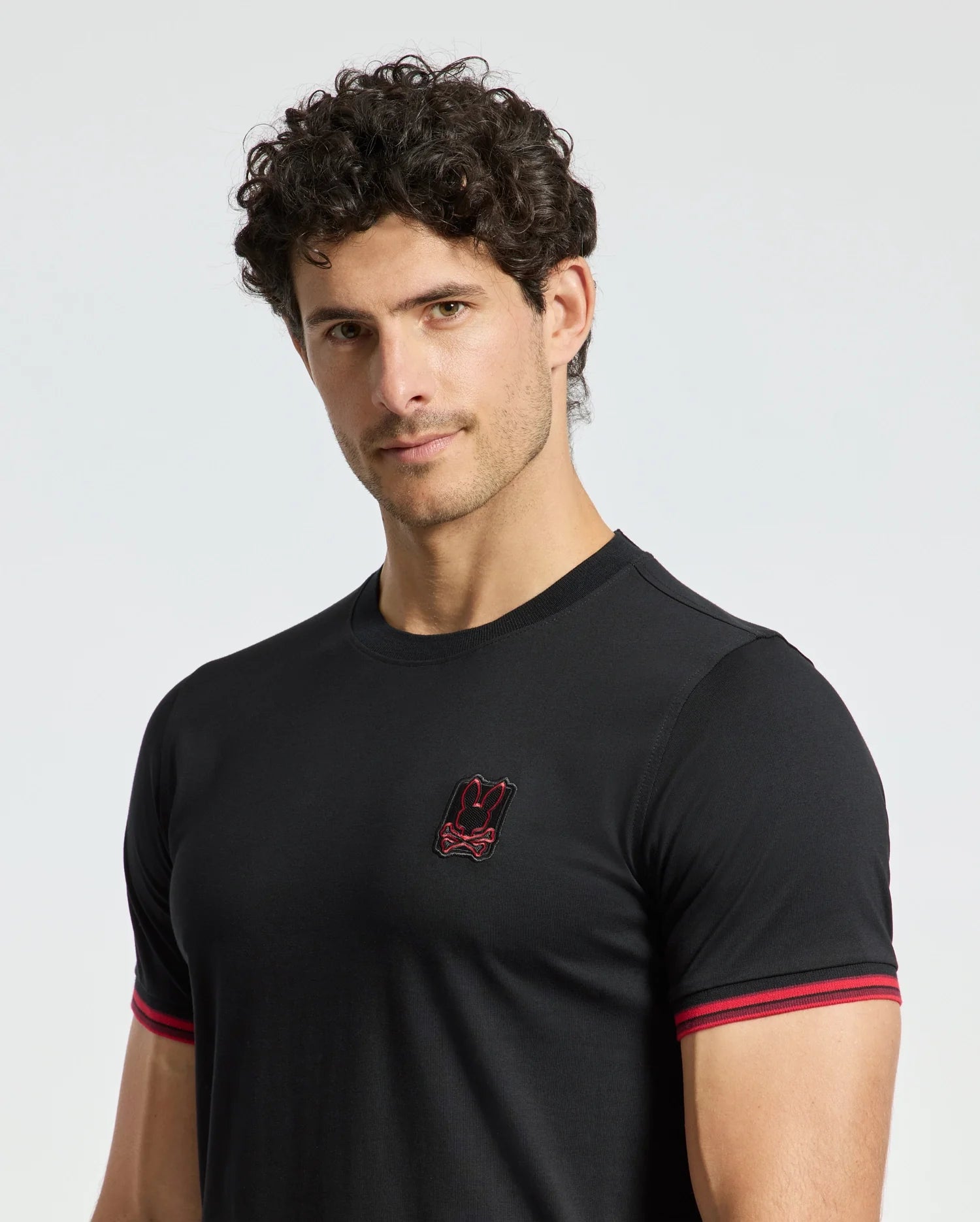 A man with curly hair wears the fitted black MENS PIERCE FASHION TEE - B6U140E200 from Psycho Bunny, featuring red trim on the sleeves and a small embroidered Bunny patch on the chest. He stands against a plain light background.