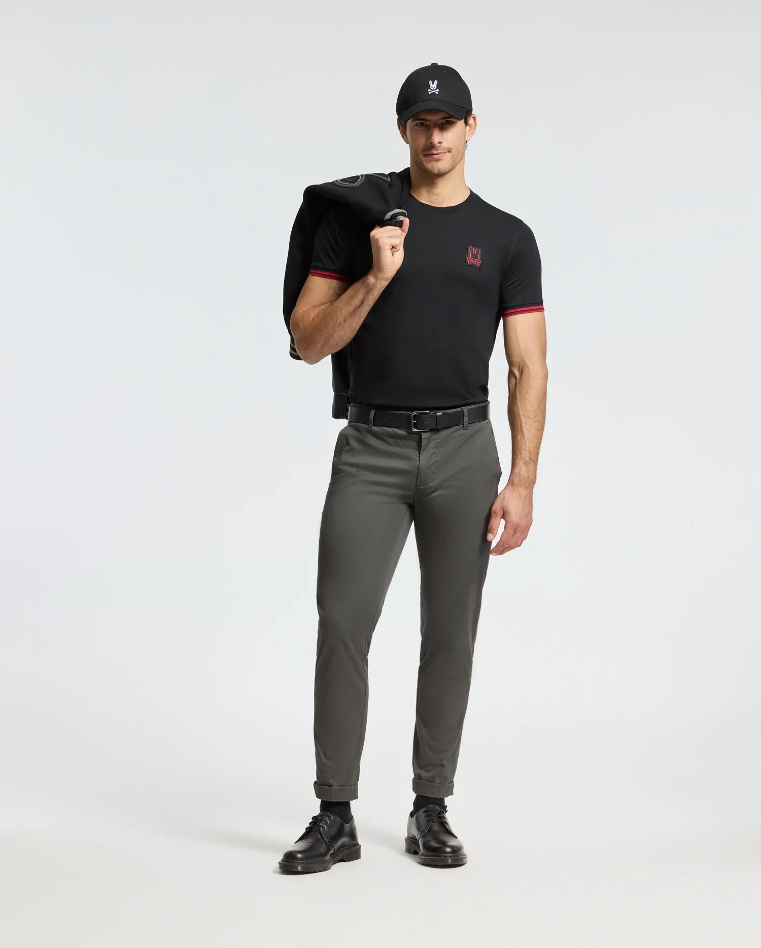 Against a plain background, a man stands confidently wearing the Psycho Bunny MENS PIERCE FASHION TEE - B6U140E200, featuring red trim and an embroidered Bunny patch. He pairs it with olive green pants, a black cap, and black shoes, effortlessly draping a jacket over his shoulder.