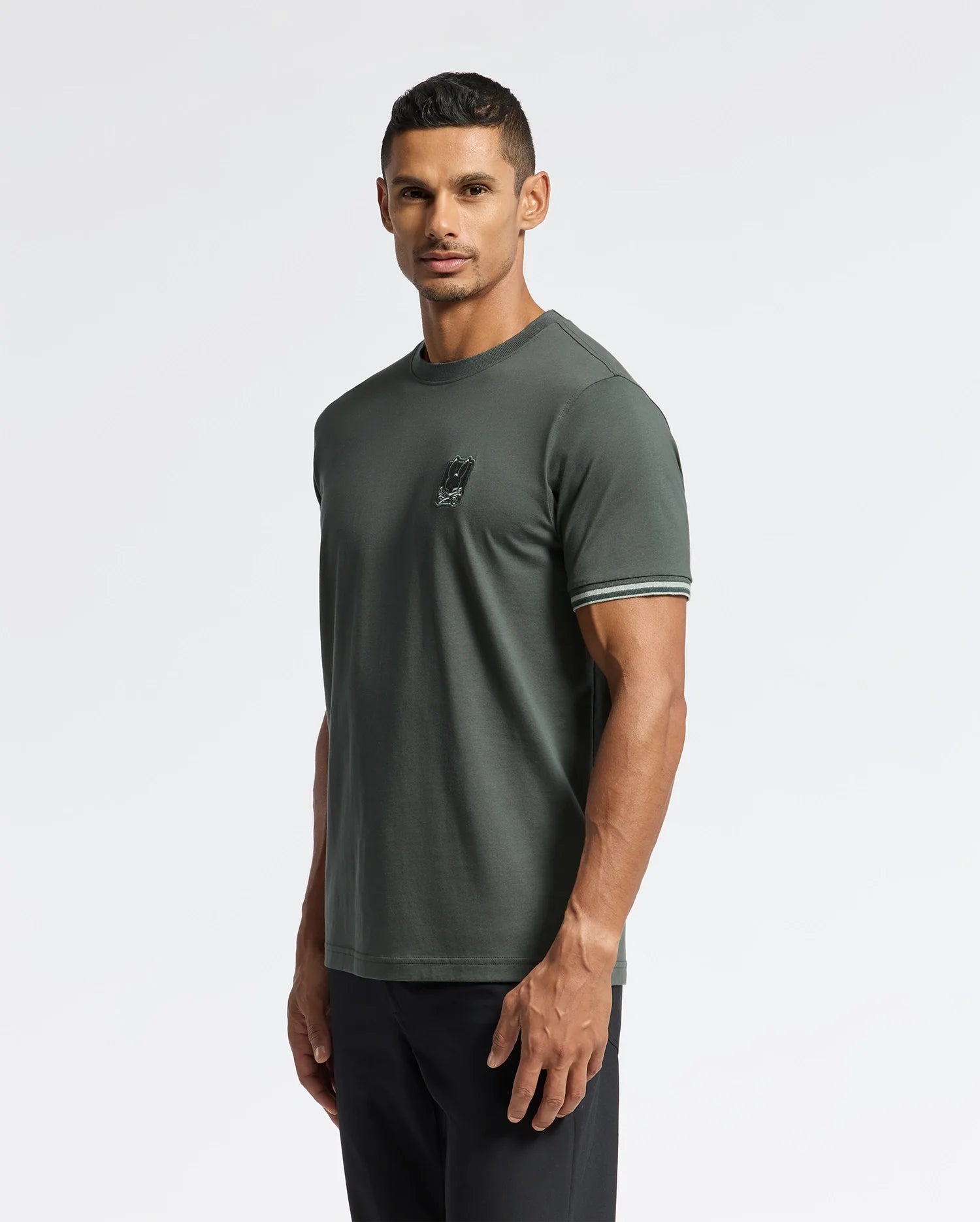 A man wearing the Psycho Bunny MENS PIERCE FASHION TEE - B6U140E200 in dark green, featuring a small logo on the chest and striped sleeve edges, stands against a plain white background. He gazes towards the camera with a neutral expression and pairs the tee effortlessly with black pants.