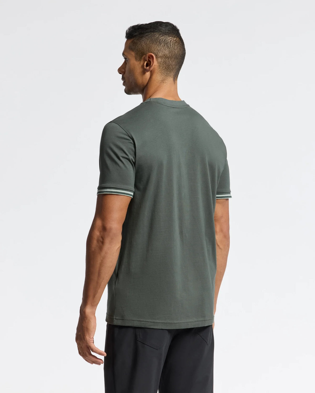 A man is standing with his back to the camera, wearing a Psycho Bunny Men's Pierce Fashion Tee in dark green featuring striped short sleeves and black pants. The background is plain white.