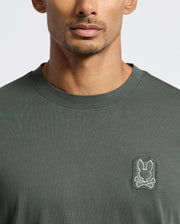 A person is wearing a dark green MENS PIERCE FASHION TEE - B6U140E200 by Psycho Bunny, crafted from soft Pima Cotton, and featuring a small embroidered skull and bunny logo on the chest. The image captures the upper torso and part of the face.