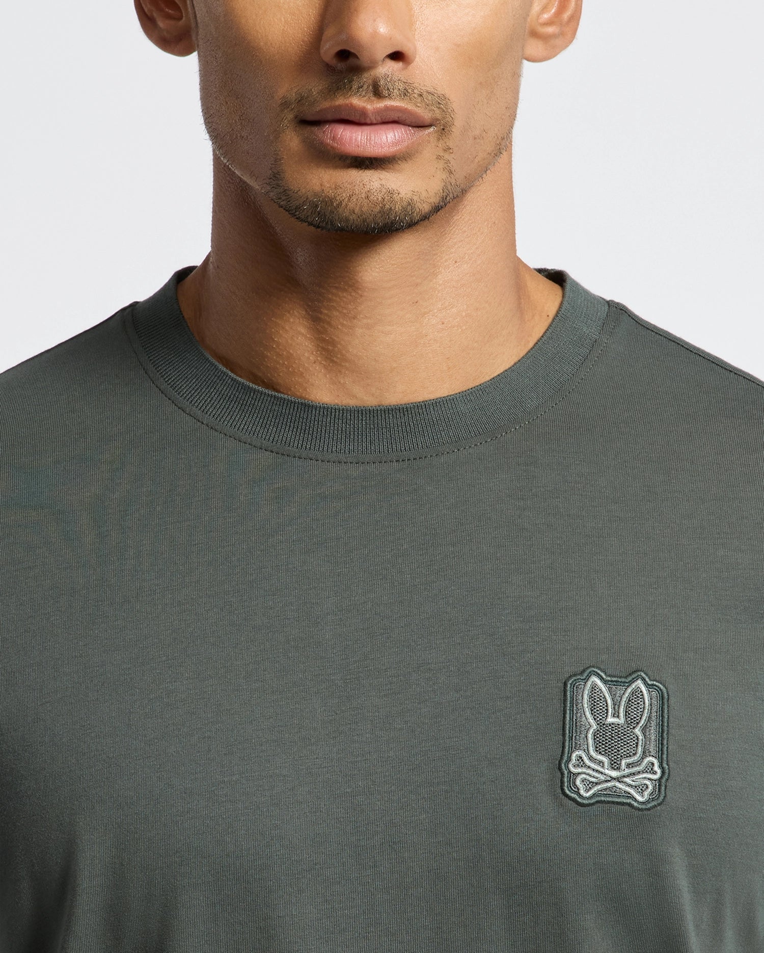 A person is wearing a dark green MENS PIERCE FASHION TEE - B6U140E200 by Psycho Bunny, crafted from soft Pima Cotton, and featuring a small embroidered skull and bunny logo on the chest. The image captures the upper torso and part of the face.