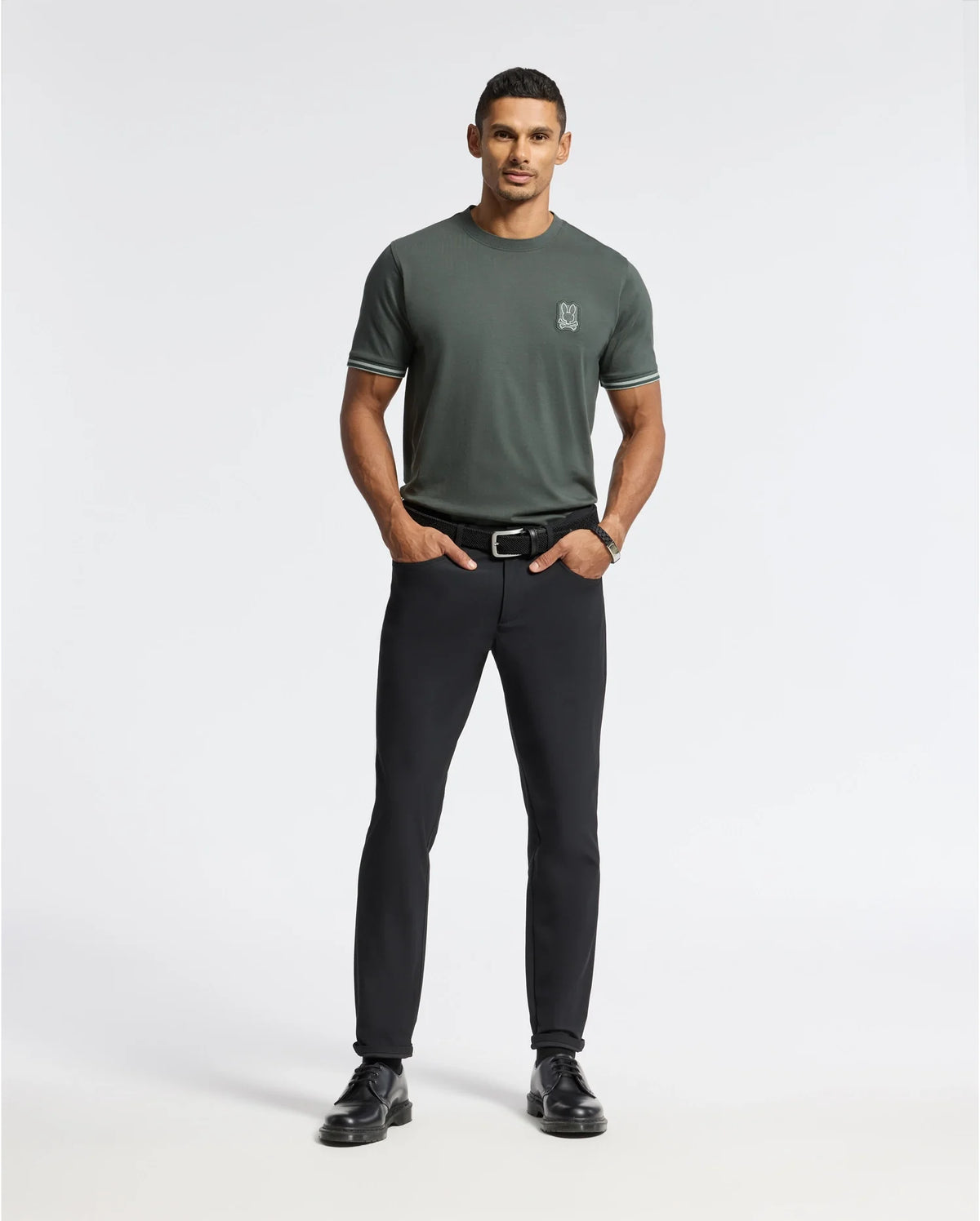 A man stands confidently against a plain white background, wearing a green MENS PIERCE FASHION TEE - B6U140E200 by Psycho Bunny, along with black pants and black shoes. His hands are in his pockets, and he has short hair and a slight smile.