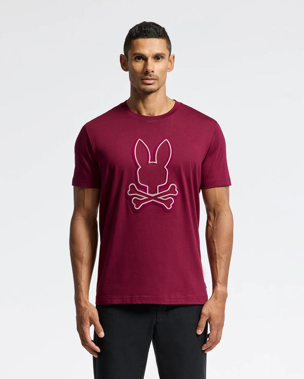 A man models the MENS ALEXANDER GRAPHIC TEE - B6U163E200 by Psycho Bunny, a stylish graphic T-shirt made from premium Pima cotton. The maroon tee showcases a white outline design of a rabbit head and crossbones. He stands against a plain white background, sporting short hair and black pants for a sleek look.