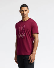 A man wearing the Psycho Bunny MENS ALEXANDER GRAPHIC TEE - B6U163E200, which features a maroon design with a rabbit's head and crossbones, stands against a plain background. Made from pima cotton, this T-shirt ensures comfort. He has short dark hair and gazes at the camera with a neutral expression.