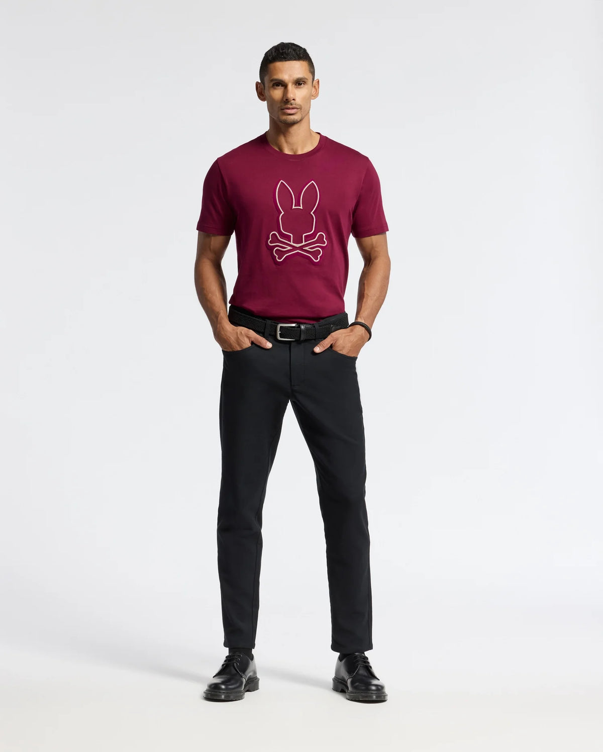 A man is standing against a simple light background, clad in the Psycho Bunny MENS ALEXANDER GRAPHIC TEE - B6U163E200, which showcases a rabbit and crossbones design in burgundy. He's also wearing black pants and shoes. His hands are tucked in his pockets, and he sports short hair.