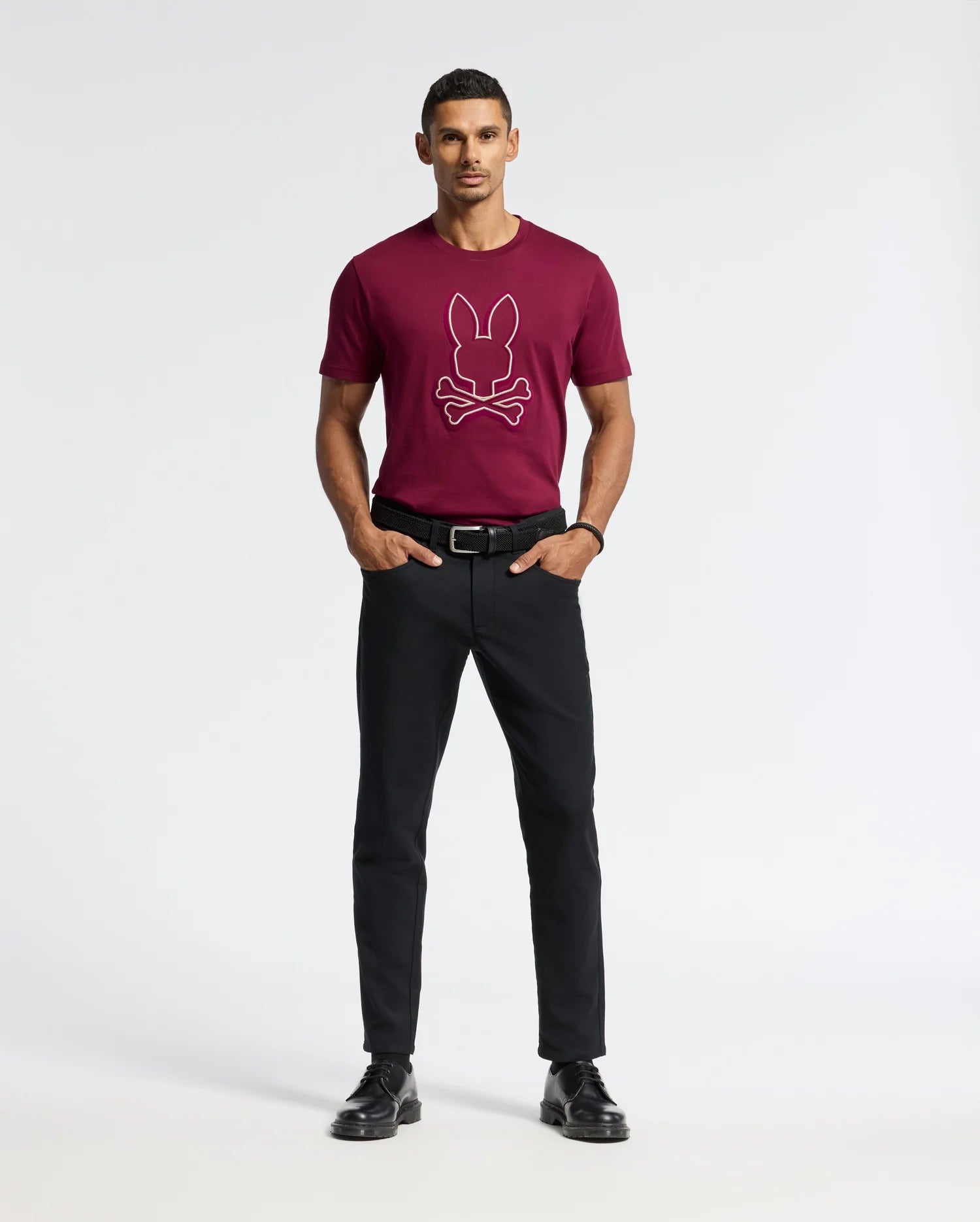 A man is standing against a simple light background, clad in the Psycho Bunny MENS ALEXANDER GRAPHIC TEE - B6U163E200, which showcases a rabbit and crossbones design in burgundy. He's also wearing black pants and shoes. His hands are tucked in his pockets, and he sports short hair.