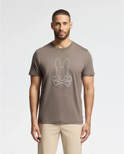 A man with short hair and a beard stands facing the camera, wearing the Psycho Bunny MENS ALEXANDER GRAPHIC TEE - B6U163E200 in brown, featuring a white outline of a rabbit and crossbones. The tee is made from soft Pima cotton. He pairs it with light beige pants, all set against a plain white background.