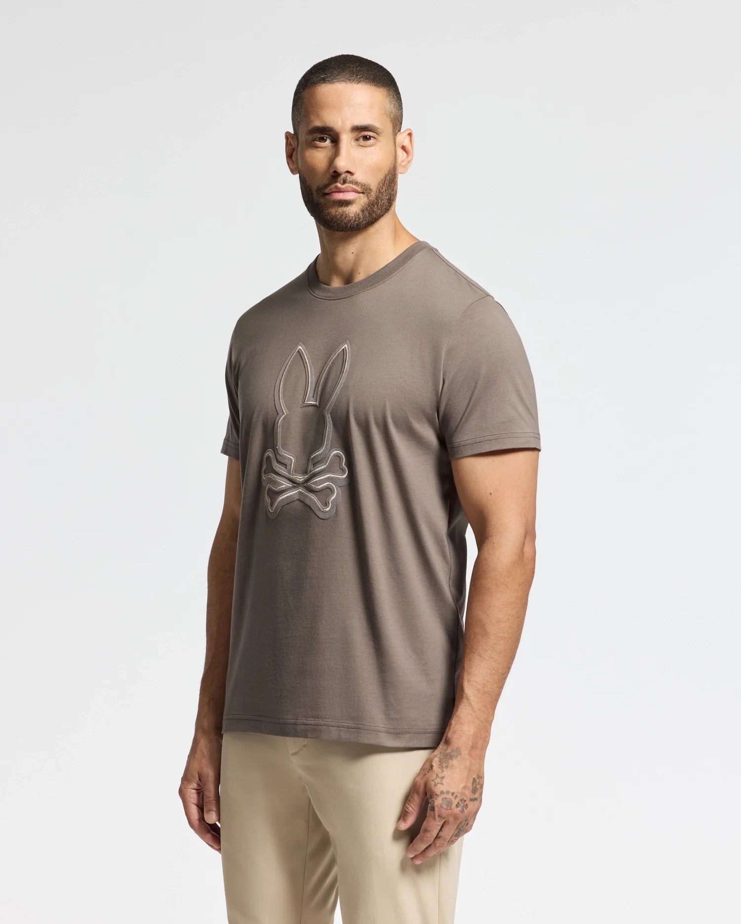 A man wearing a taupe Psycho Bunny Men's Alexander Graphic Tee, with a bunny and crossbones design, stands against a plain backdrop. He is also dressed in light-colored pants and has tattoos on his left hand and forearm.
