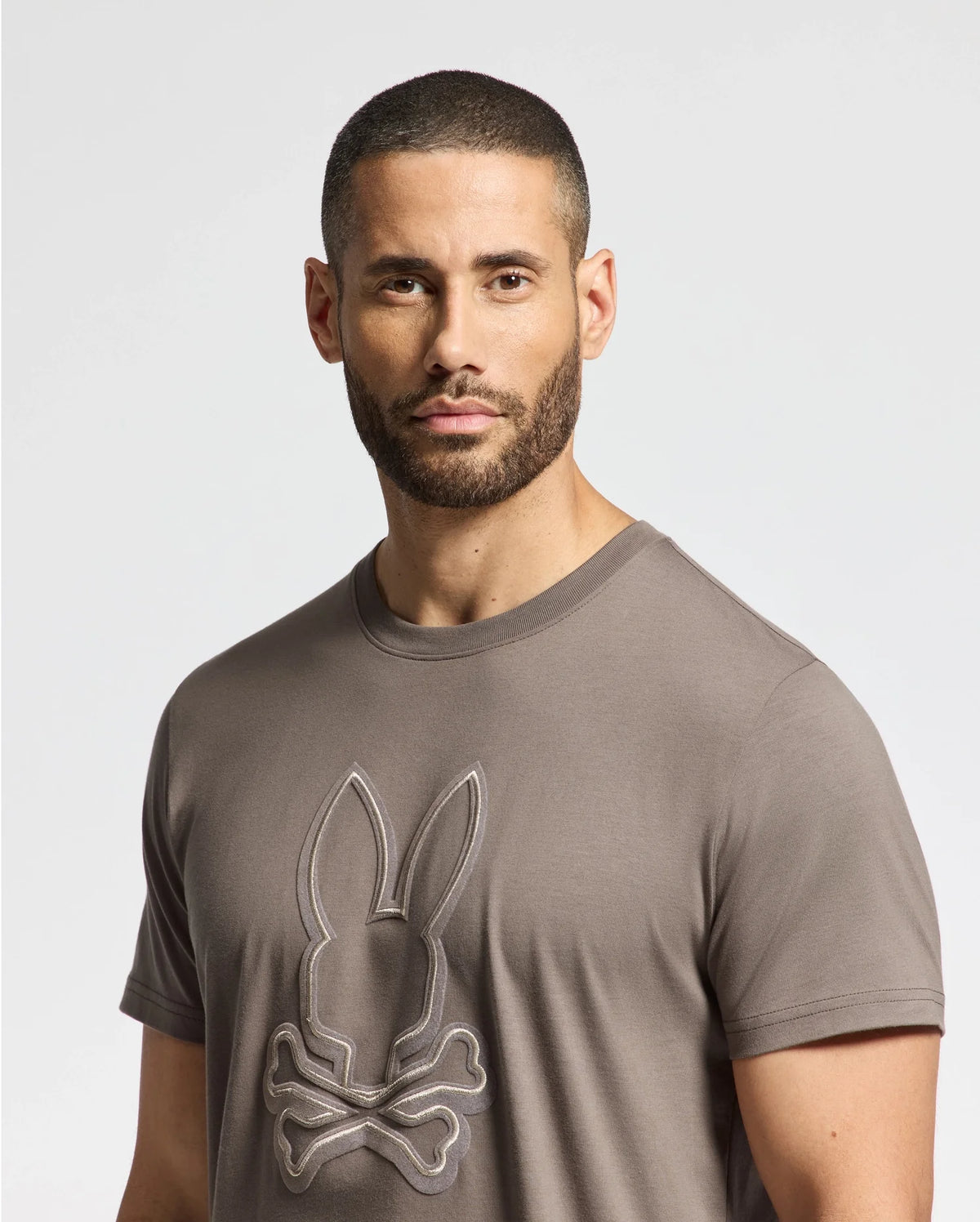 A man with short hair and a beard is wearing the Psycho Bunny Men's Alexander Graphic Tee, crafted from soft Pima cotton. The gray shirt features a stylish rabbit and crossbones design with subtle Lurex details, as he stands against a plain white background, looking directly at the camera.