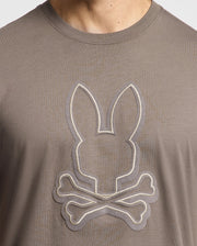 A person wearing the Psycho Bunny MENS ALEXANDER GRAPHIC TEE - B6U163E200, crafted from soft pima cotton, showcases an embroidered bunny head above crossed bones for a playful take on the traditional skull and crossbones design.