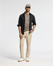 A man stands against a plain background, wearing a gray cap and an open black shirt. His outfit showcases the MENS ALEXANDER GRAPHIC TEE - B6U163E200 by Psycho Bunny, crafted from soft Pima cotton, paired with beige pants and white sneakers.